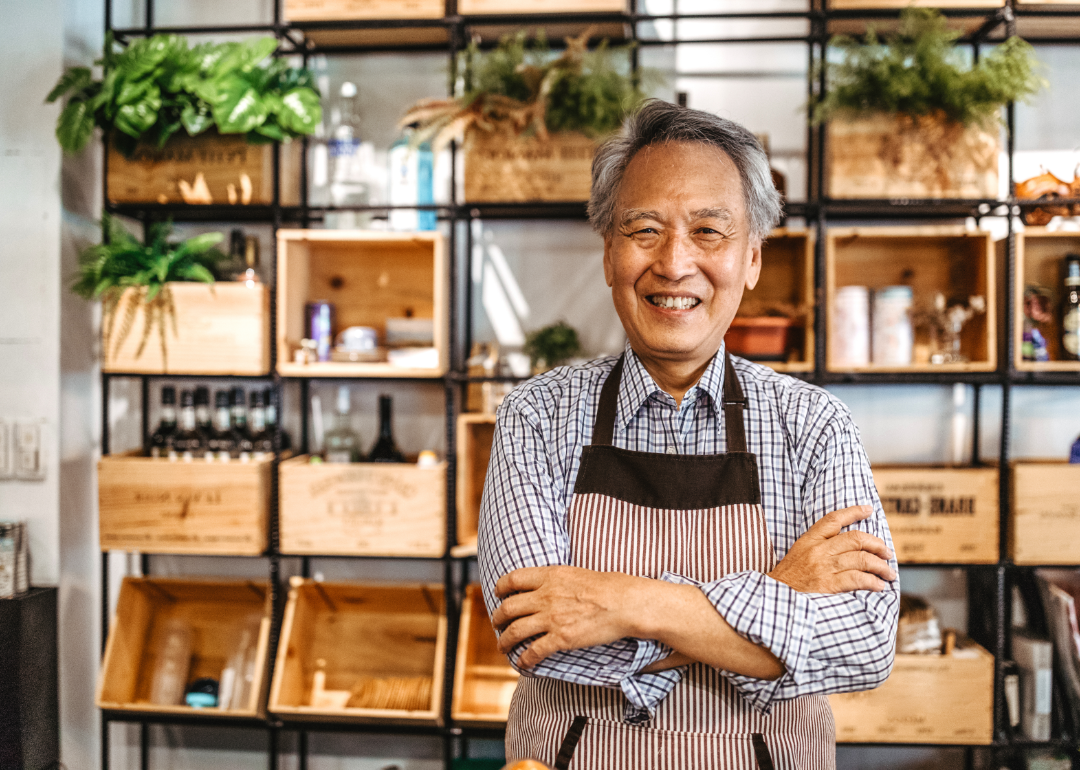 An older man smiling with his arms folded in a shop with shelves behind him.