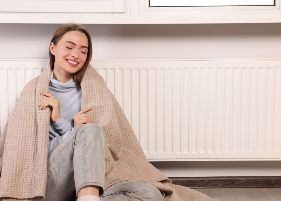 A young woman sits on the floor, wrapped in a blanket, near a wall radiator unit