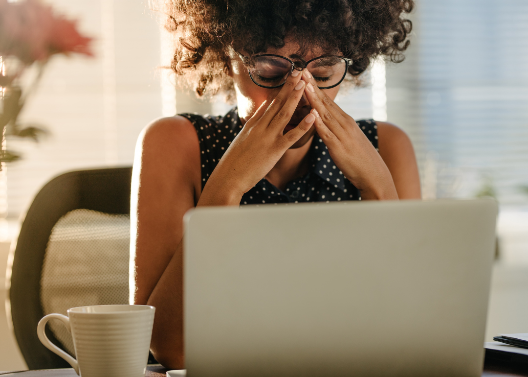 A Black woman sits in front of a laptop in a stressed posture, her hands covering part of her face