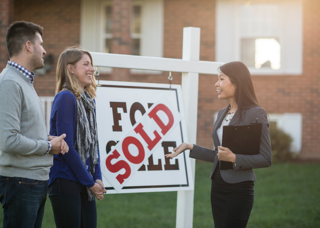 A couple stand in front of a fore sale sign with a 'sold' sign pasted into it, speaking with a realtor