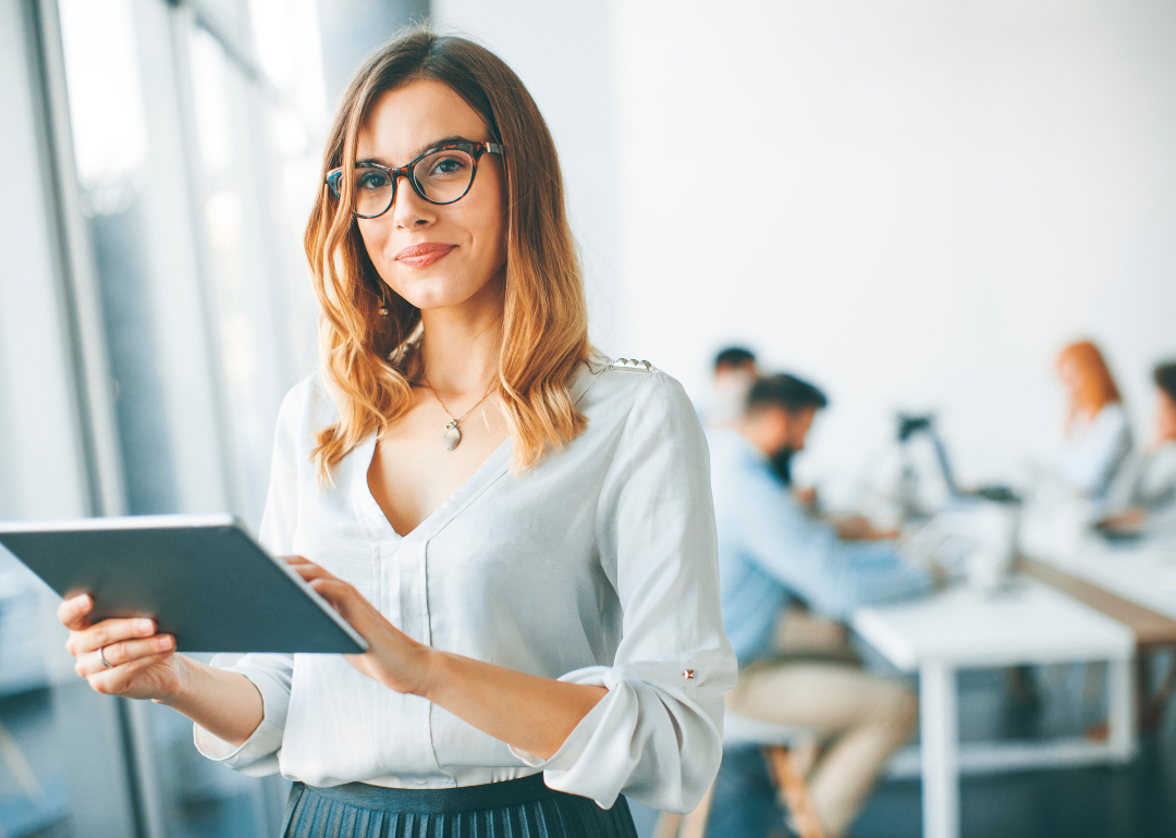 A young female professional wearing glasses holds a tablet in an office setting