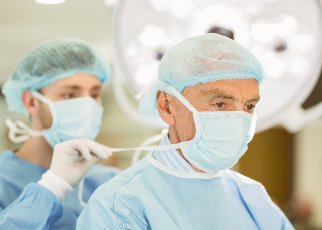 A younger nurse help secure an older surgeon's mask.
