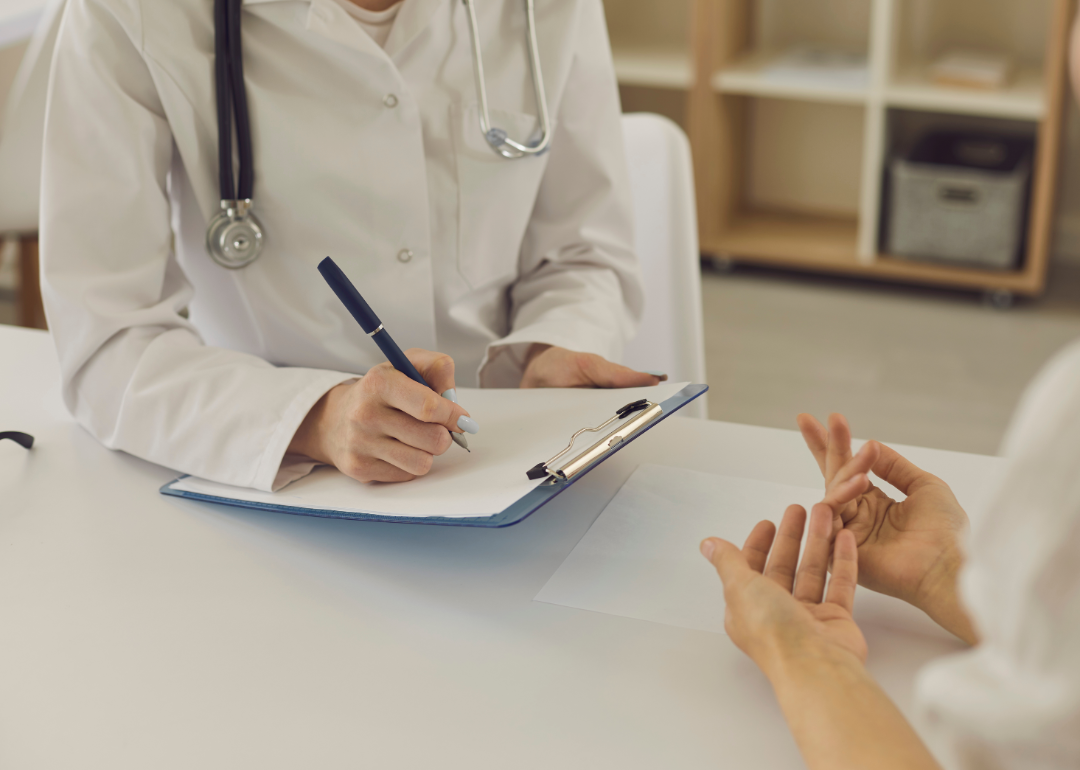 A doctor writes on a clipboard as they speak with a patient at a desk.