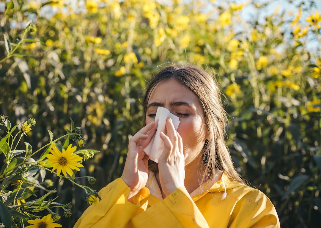 Person sneezes into a tissue due to allergies while surrounded by flowers.