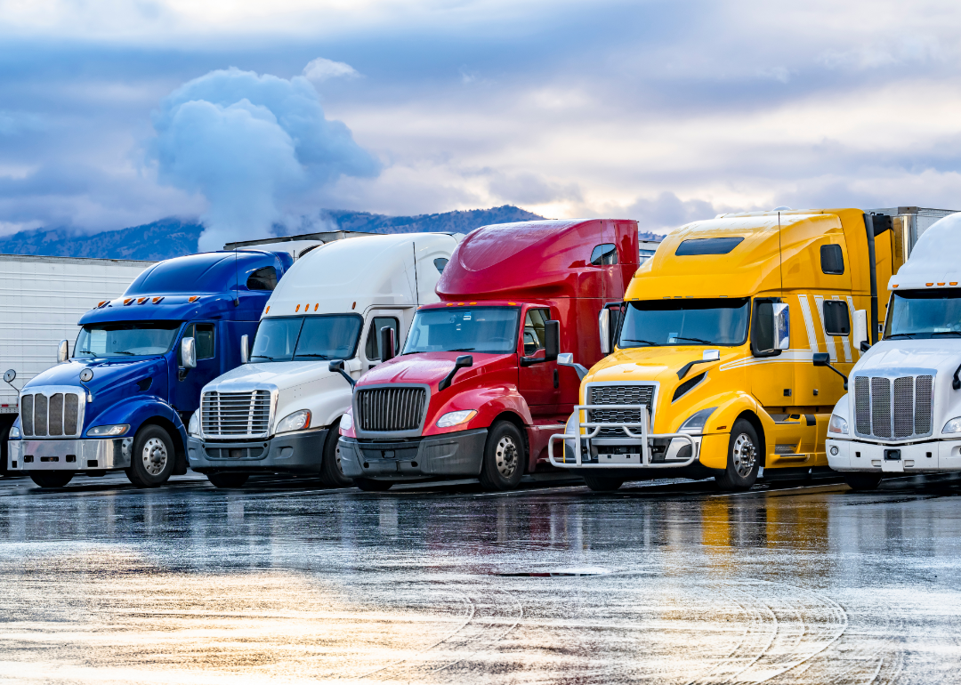 Colorful big rig semi trucks with semi trailers parked closely in a lot.