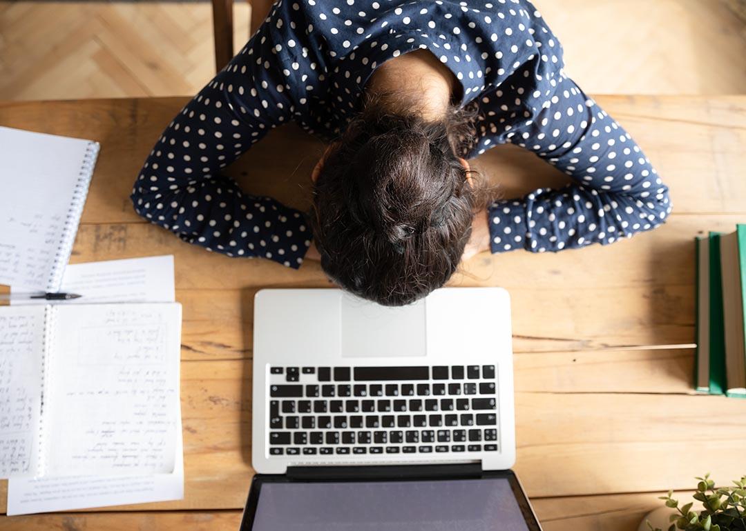 overhead view of woman resting head on desk in front of laptop computer and notes