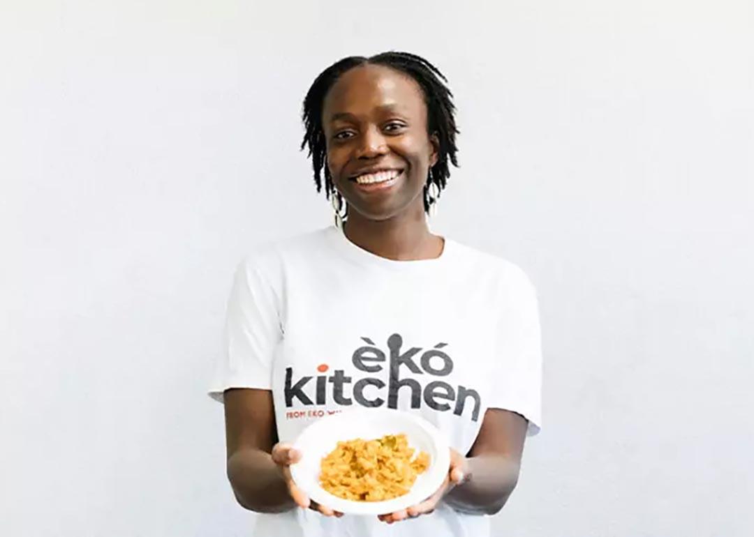 Simileoluwa Adebajo wears a shirt from her business Eko Kitchen and holds a plate of food, standing in front of white background.
