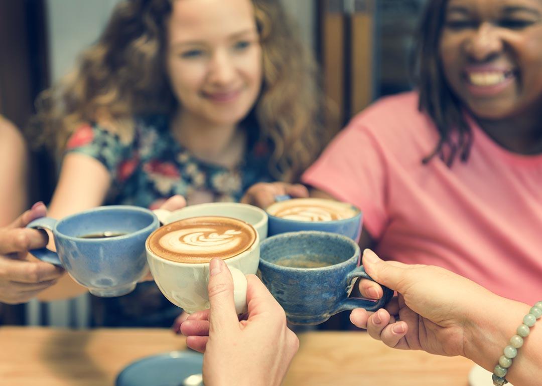 Five hands holding coffee touch their drinks together in the middle of the table with two women's face visible in background.
