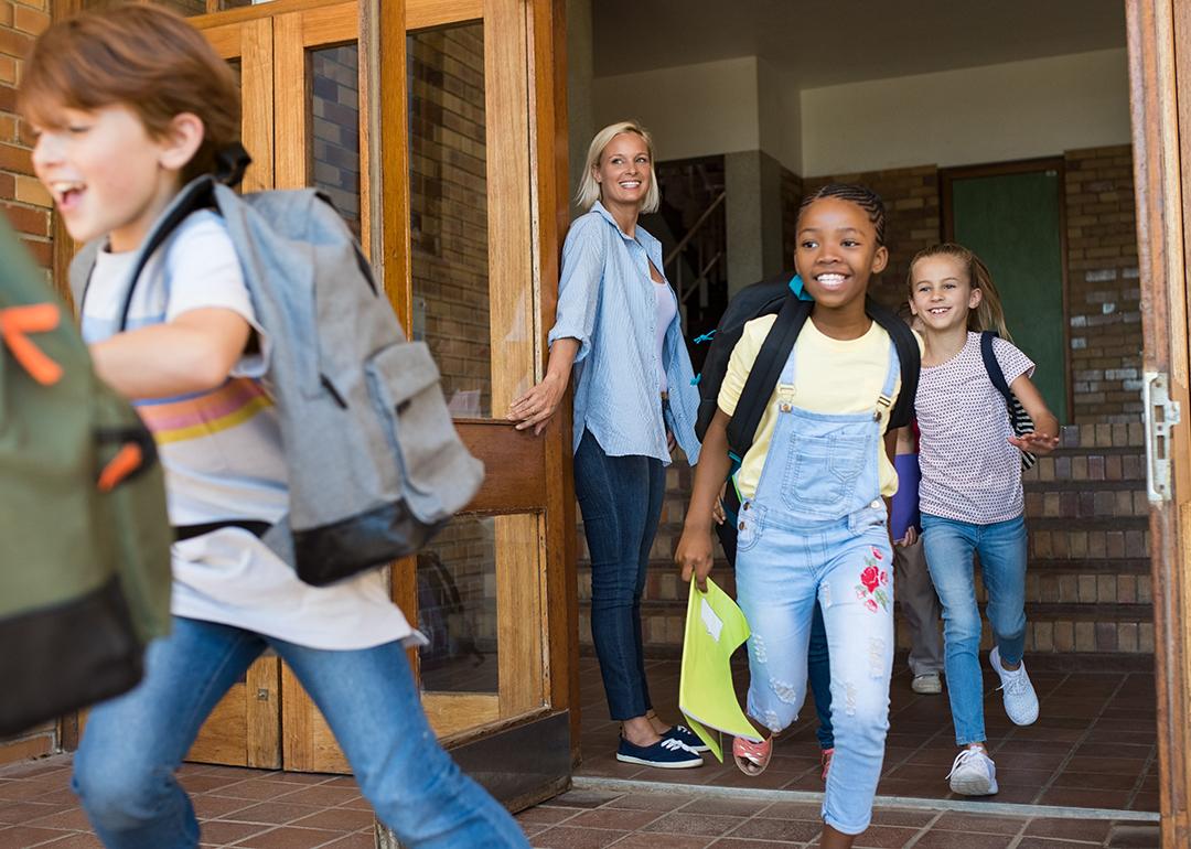 Smiling elementary school-age students run out of a school from a door held open by a teacher.