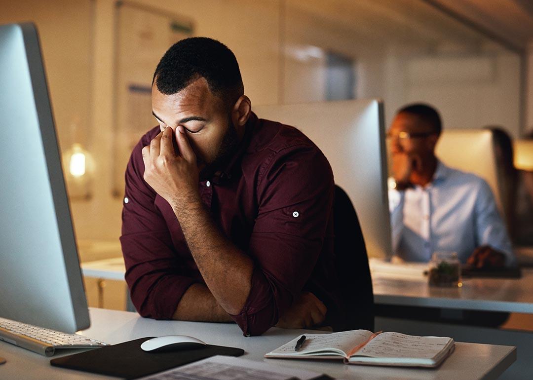 A man in front of a computer at a desk showing a burnout or headache gesture.