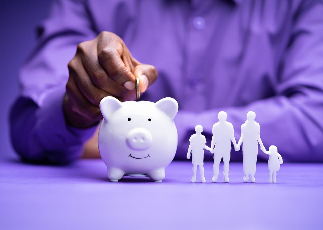 A focus on a person's hand while dropping a coin in a piggy bank with a 3D figure of a 4-member family beside it.