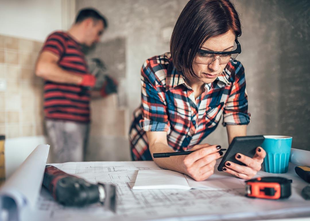 Two people in charge of overseeing a home improvement where one woman is leaning on a table with blueprints and noting with an instrument and the other is a man hammering nails on a wall.