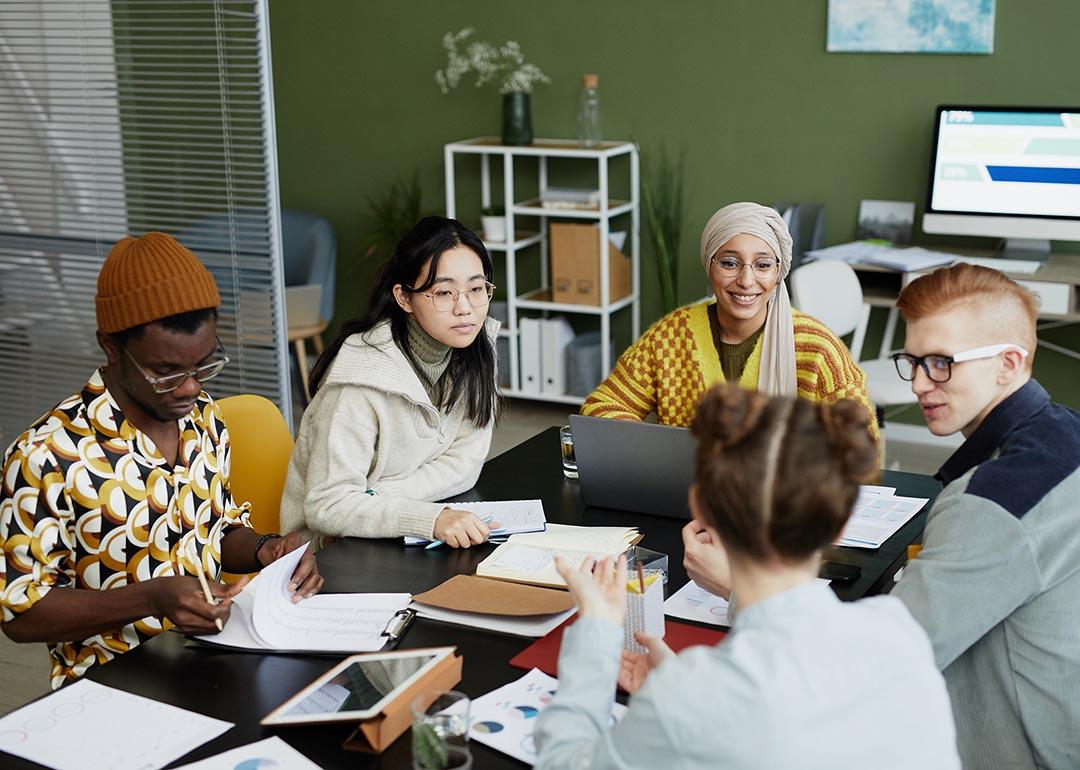 In an office, a diverse group of five young adults seated in a round table engaging in a discussion.