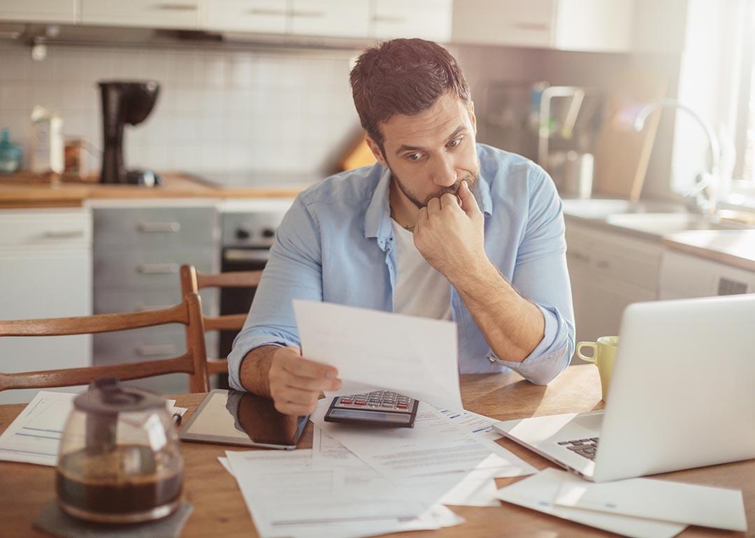 A man sitting in a kitchen with a laptop and calculator on the table while reading a letter with a focused gesture.