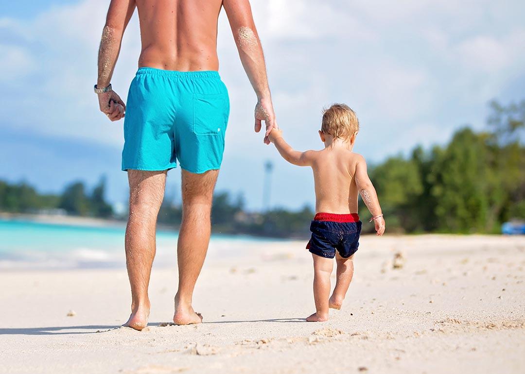 A dad and holding his toddler son's hand while they are walking on a white sand beach.