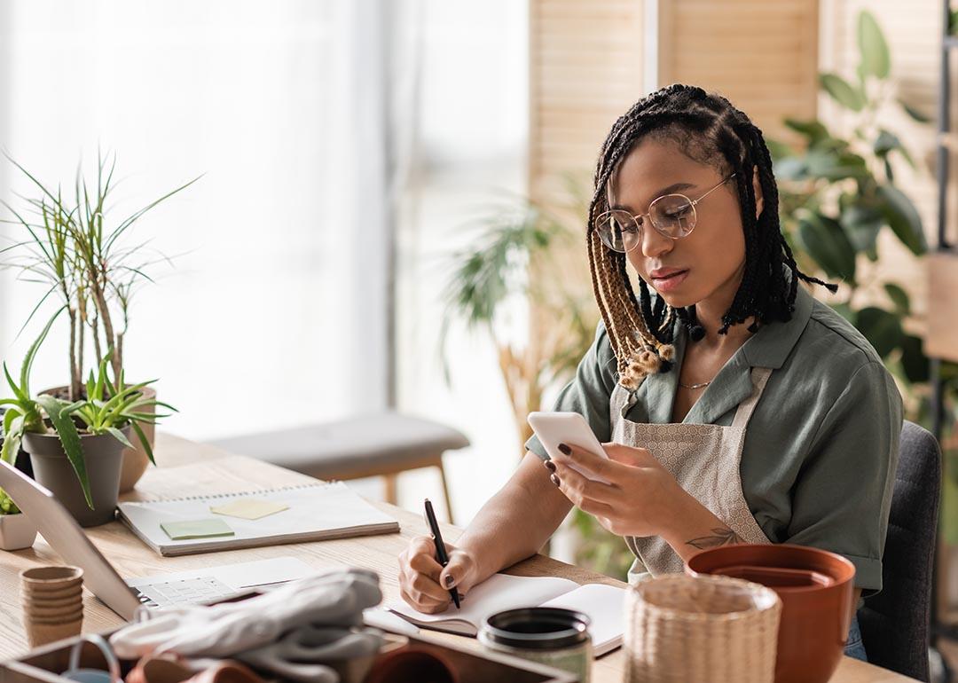 A woman with a gardening business is looking into a phone while writing down information on a notebook.