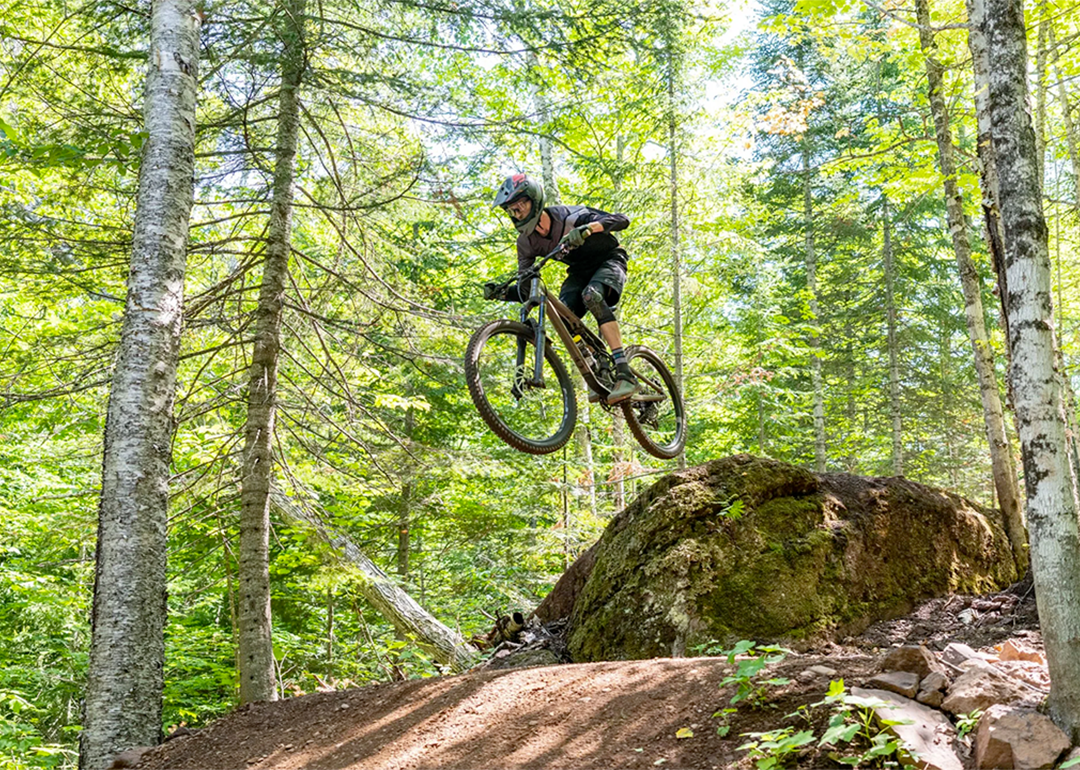Mountain biker going off a jump on a dirt track with trees in the background.