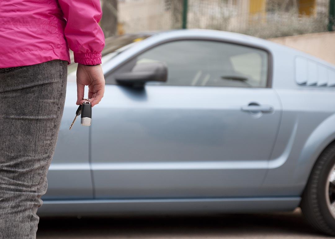 Person in dark pink shirt and black jeans holding car keys seen from behind, standing in front of used light blue car.