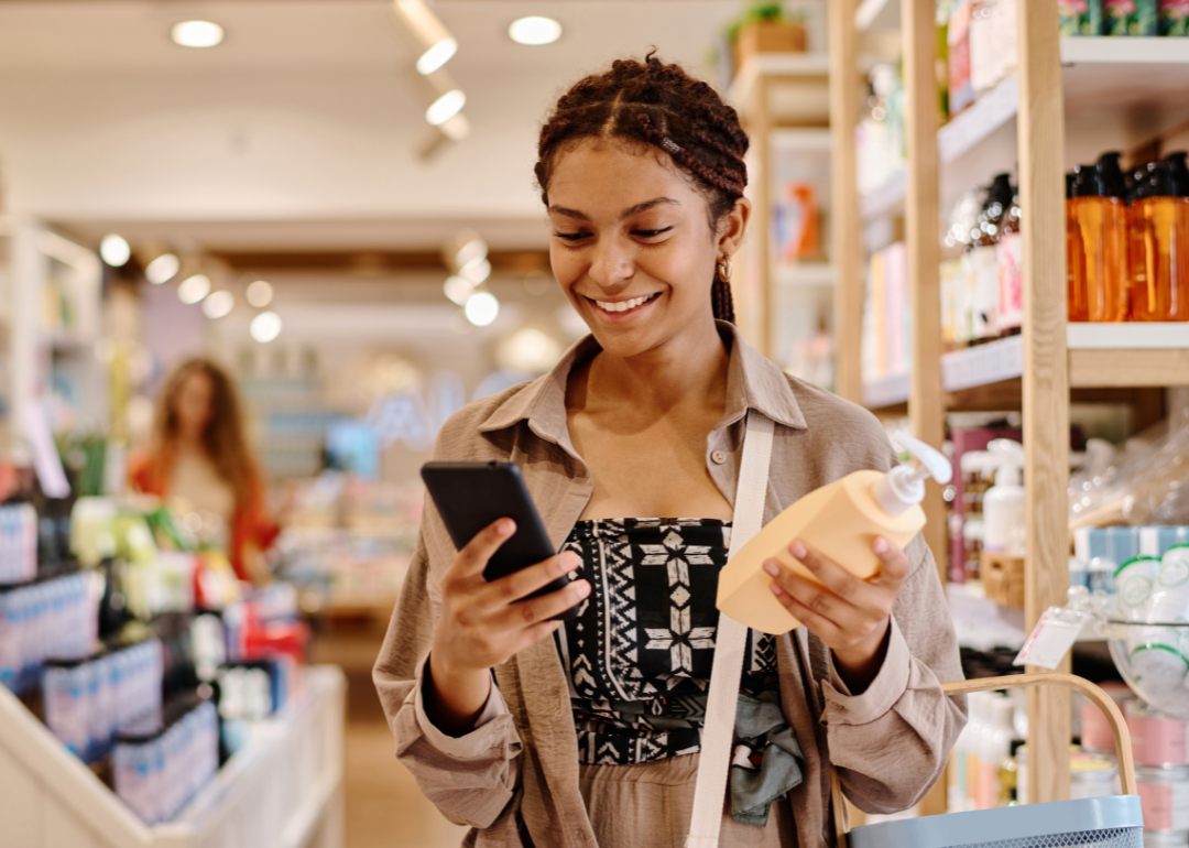 A smiling woman in a store, holding a bottle with a pump and looking at her phone.