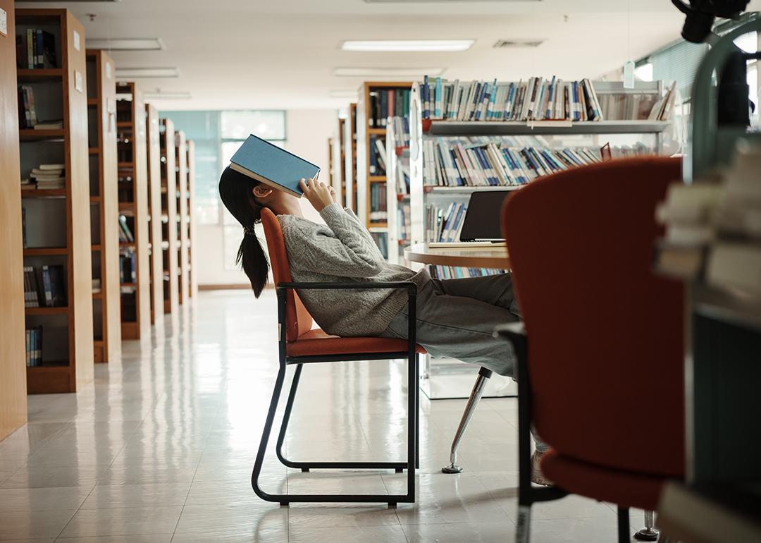A female college student in a library with an open book over her face seemingly tired of reading.