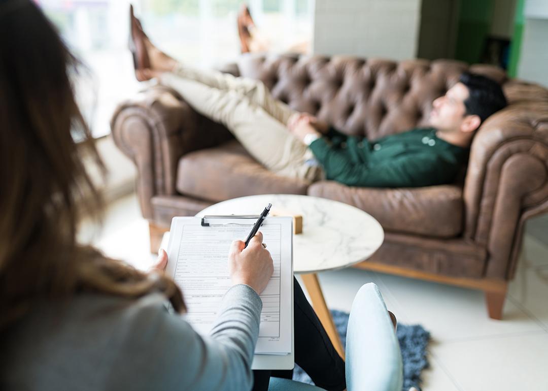 A psychotherapist writing down in a notebook while in session with a male patient who is lying on couch.