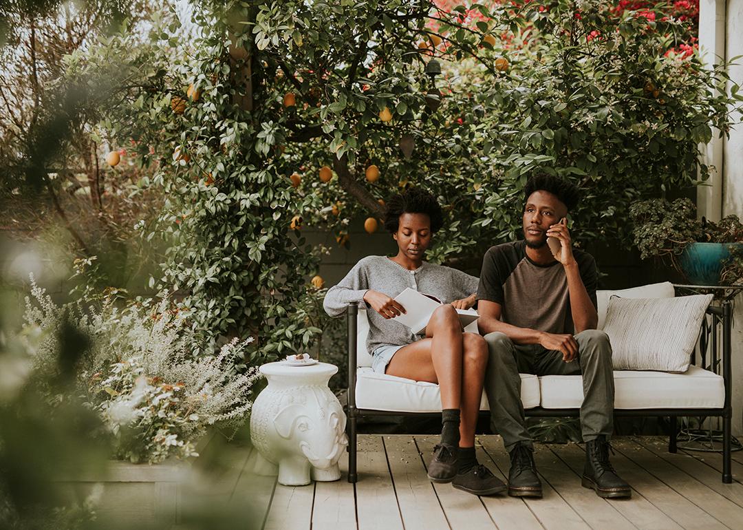 A black couple sitting on a bench in their home garden while reading a book and using a phone.