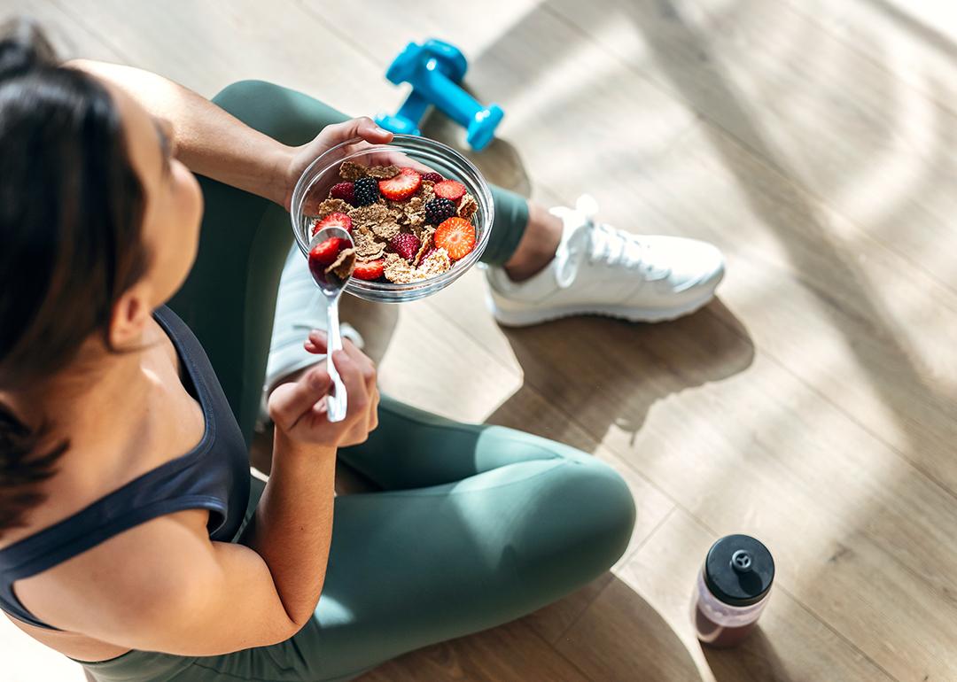A young woman sitting on the floor post-workout and eating a bowl of oatmeal with fruits.