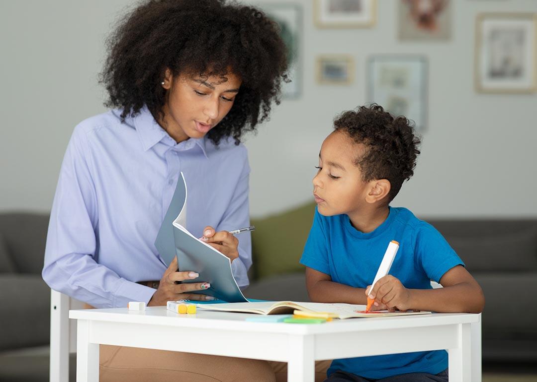 Black adult helping child learn and write, looking at a book.