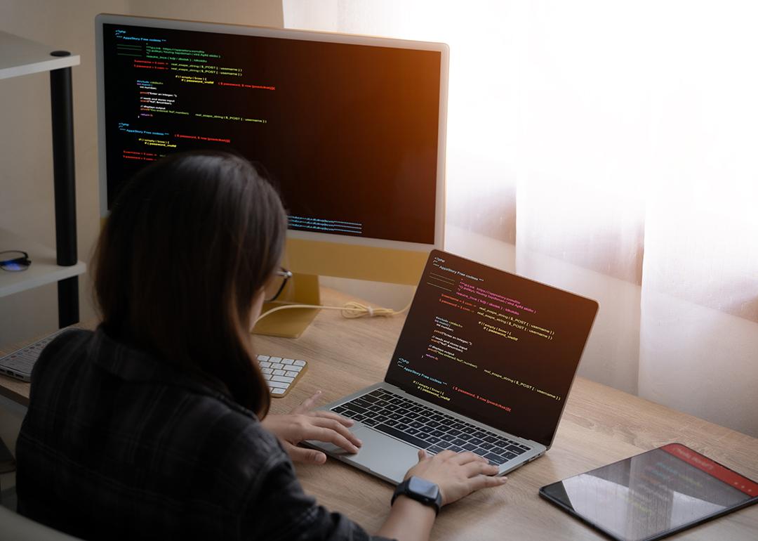 A woman working from home with a laptop and separate monitor equipment showing tech-related interface.