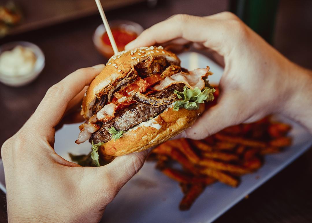 A bacon burger being held with a plate of fries in the background.