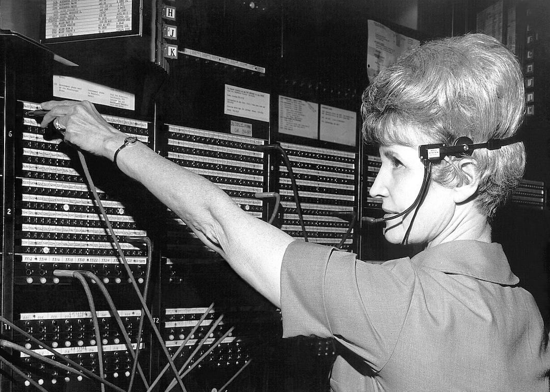 Female CDC telephone operator seated at a switchboard wearing a traditional headset, reaching for a telephone patch cable, 1974.