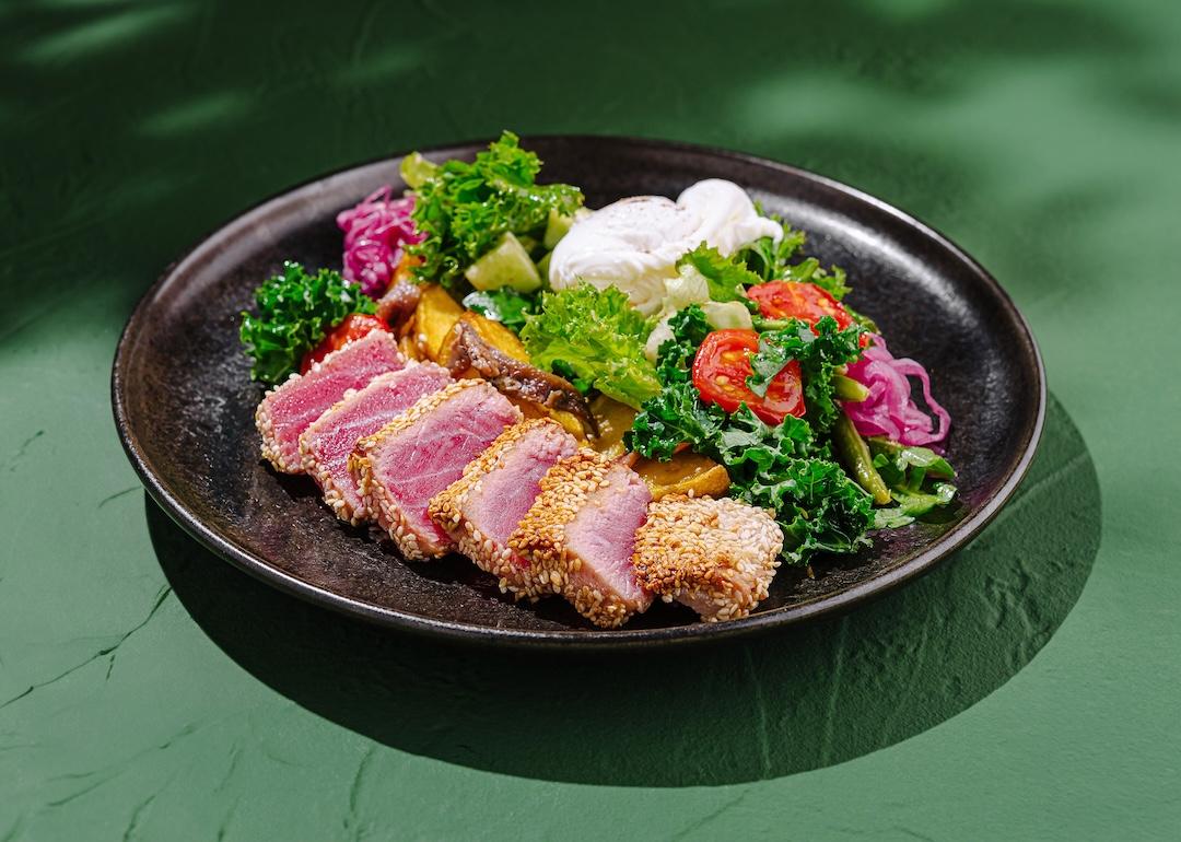 Seared tuna, coated in sesame seeds, is served on a black plate with fresh greens, colorful vegetables, and poached eggs, highlighting vibrant flavors and textures.