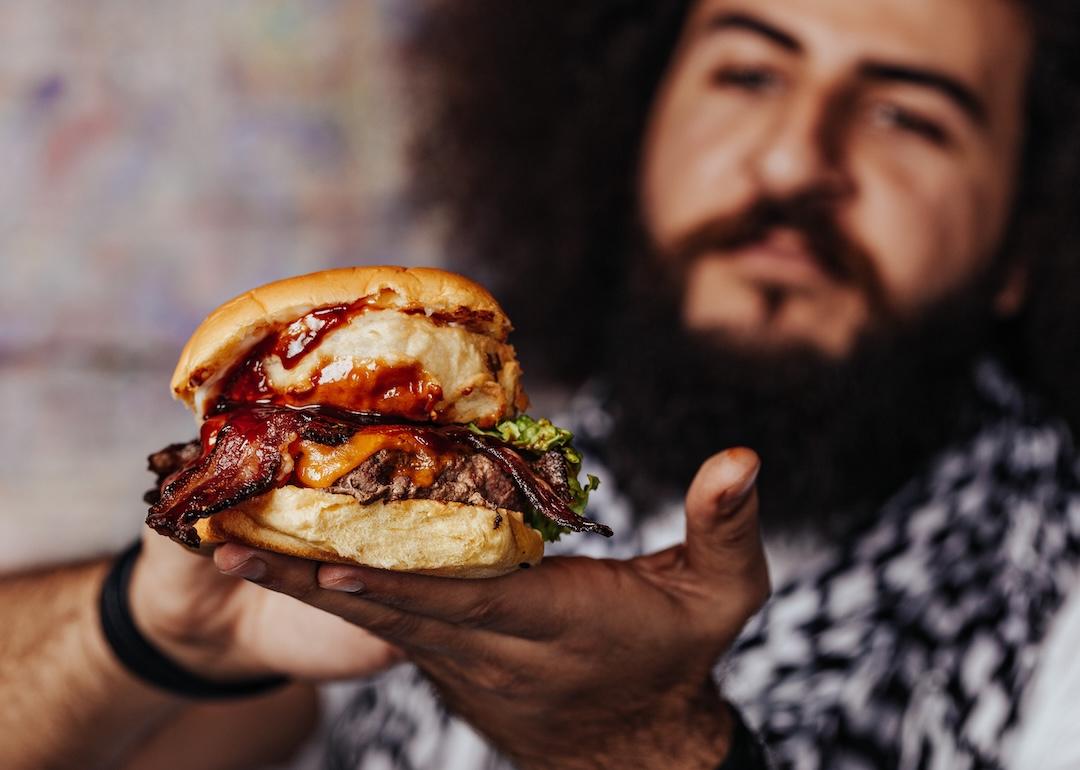 Curly hipster holding craft mouth-watering bacon and angus beef burger.
