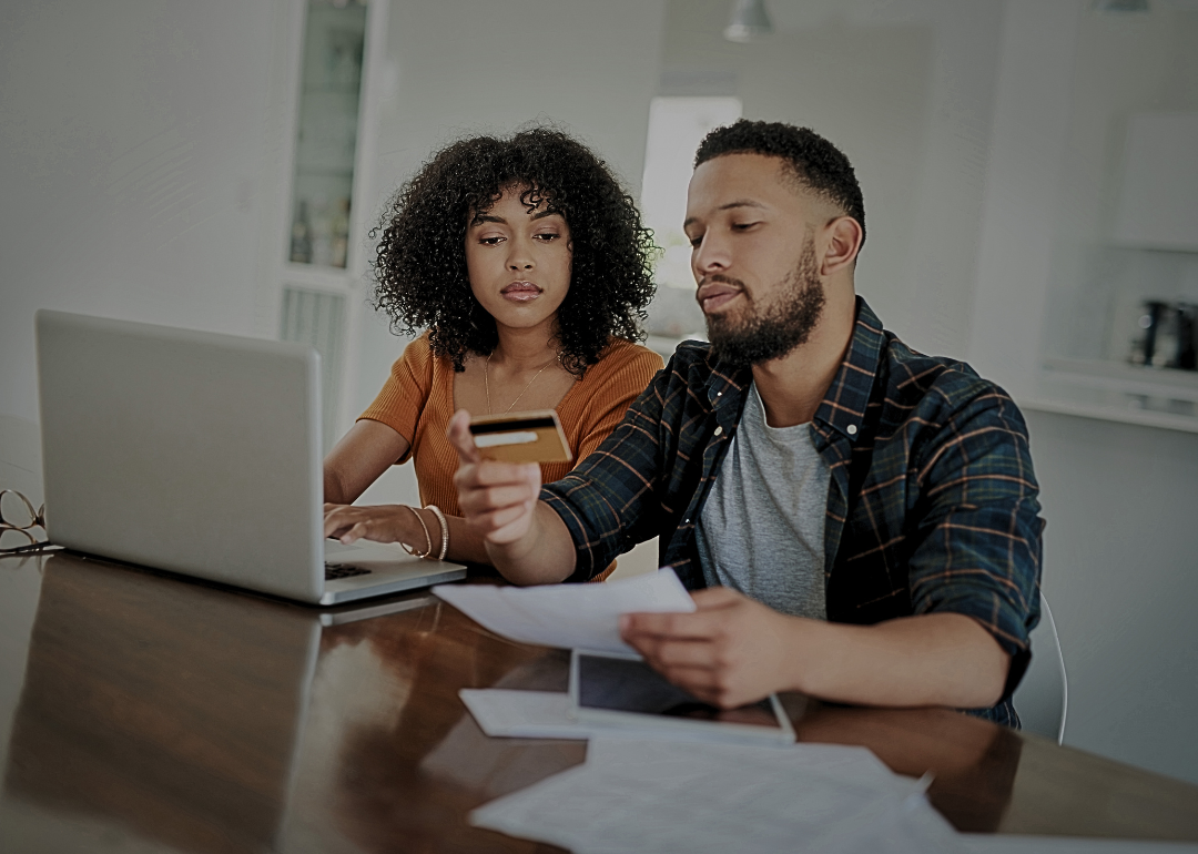 A young couple sitting at the table with a laptop, papers, and a credit card in front of them.
