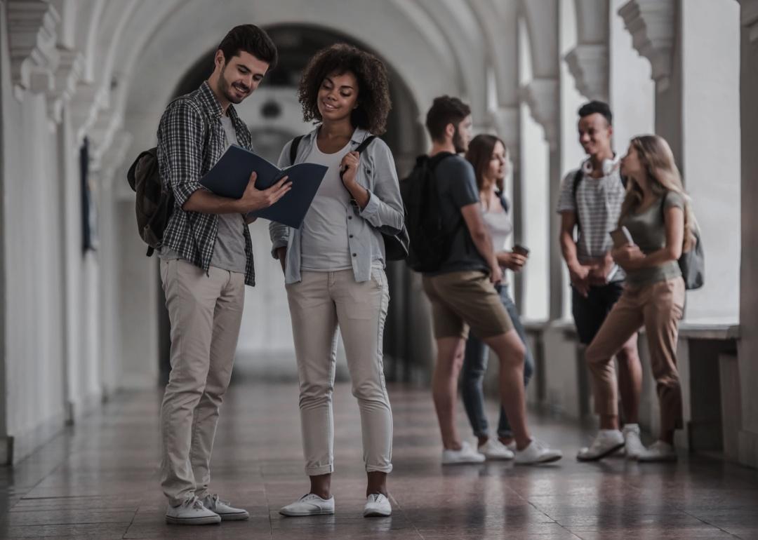 Two individuals, a man and a woman, are standing in hallway with an elegant architectural looking at a document or folder. Both are casually dressed and carry backpacks, In the background, other students are seen in small groups chatting.