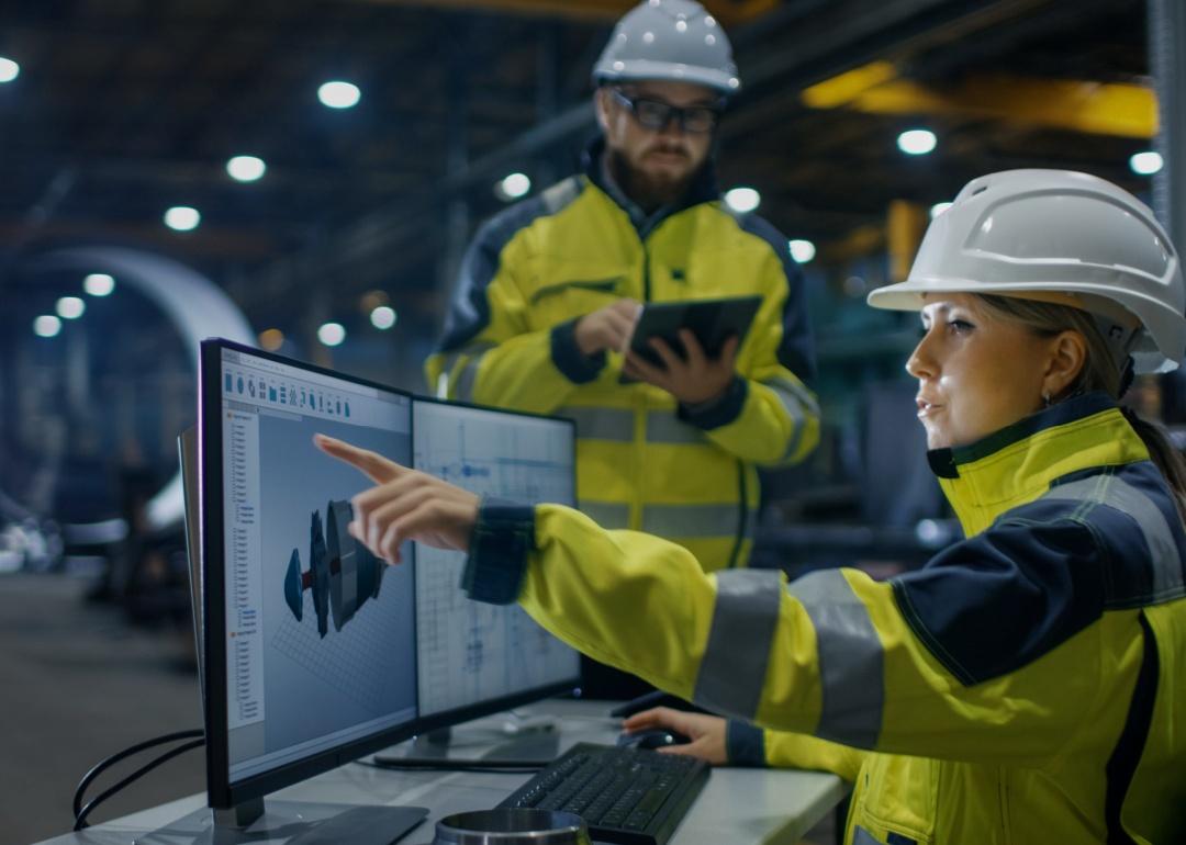 A woman construction worker wearing a heavy-duty bright green jacket and helmet sitting in front of the computer screen showing a 3D model pointing to the screen in front of a male construction worker.