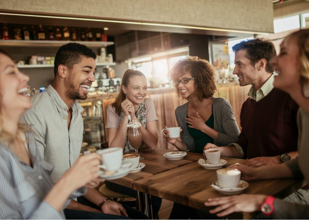 A group of six friends gathered around a wooden table in a cozy café. They are all smiling and laughing. Each person is holding a cup of coffee.