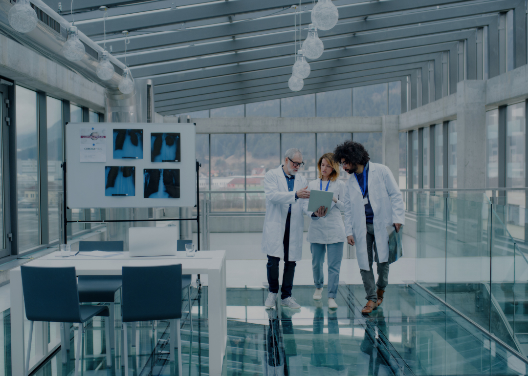 Three people in white scrubs walking down the hall of a glass, modern building looking at the document in front of them.