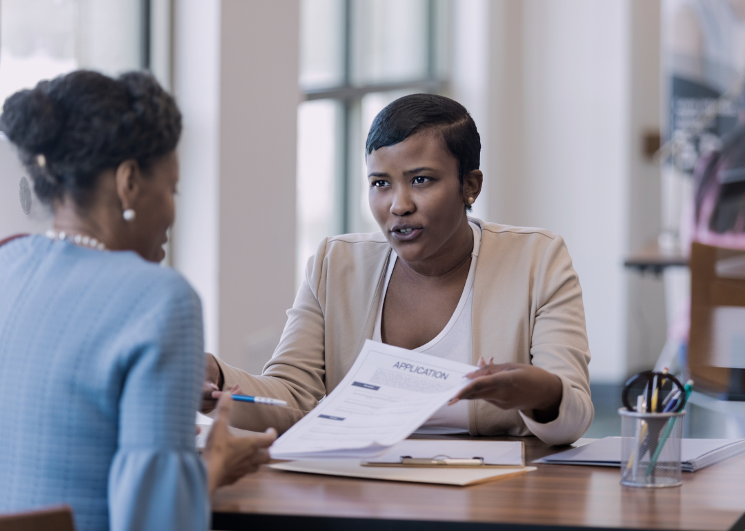 A Black woman at the desk is showing an application document to an African American woman sitting opposite her.
