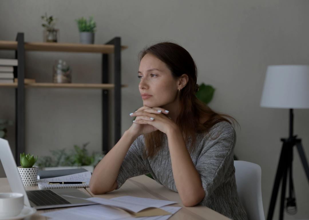 A woman sitting at a desk resting her chin on her hands with her fingers interlocked. She's looking forward. 