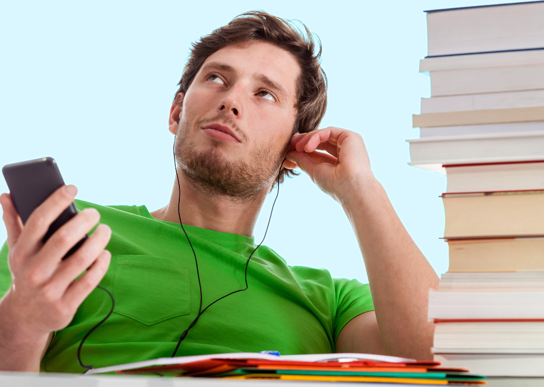 A young man sitting at the desk with a tall stack of books listining to music on his phone looking lost in his music.