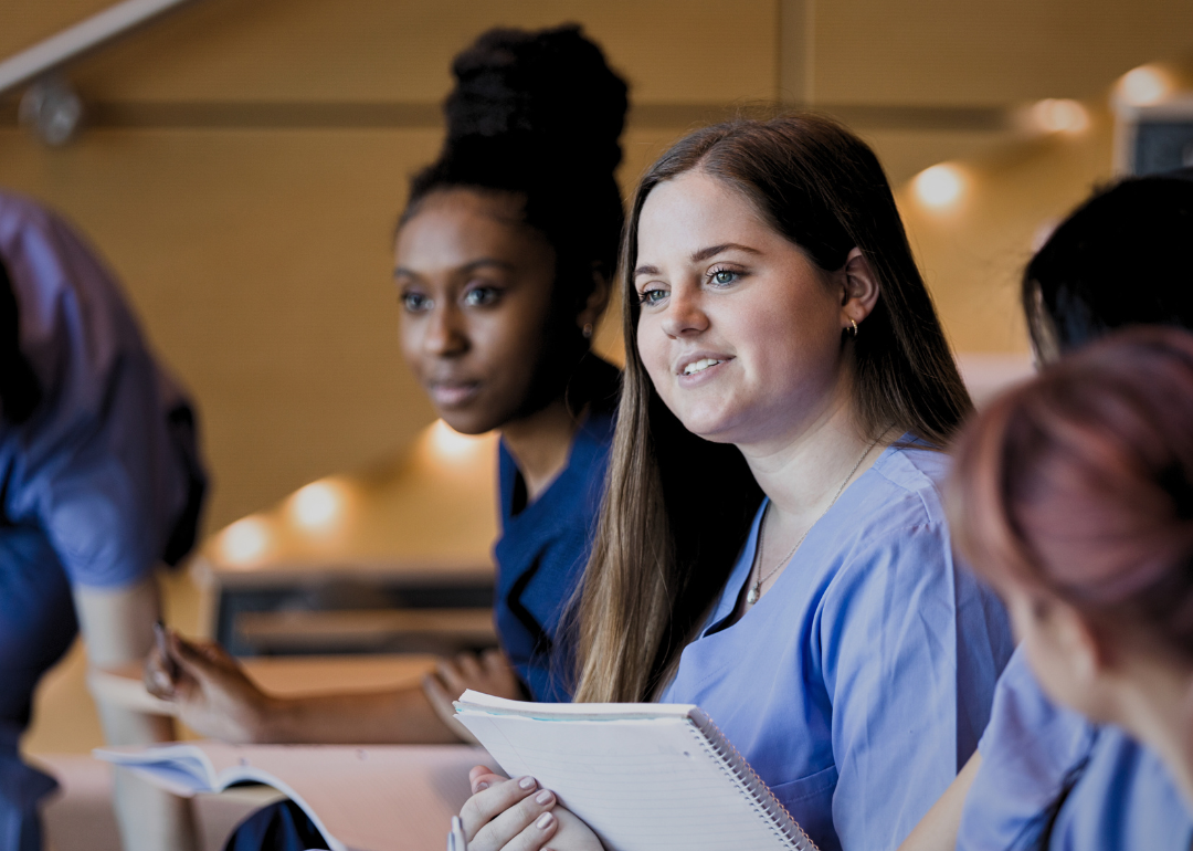 A young female dressed in blue scrubs sitting in the classroom holding a notebook looking forward. 