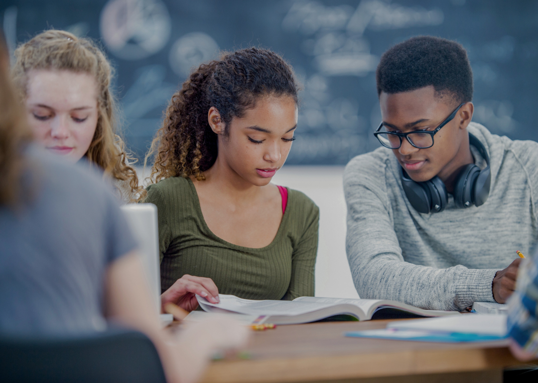 A group of diverse students sitting together in a classroom looking at the books in fornt of them. In the background, a chalkboard with various mathematical equations or diagrams visible.
