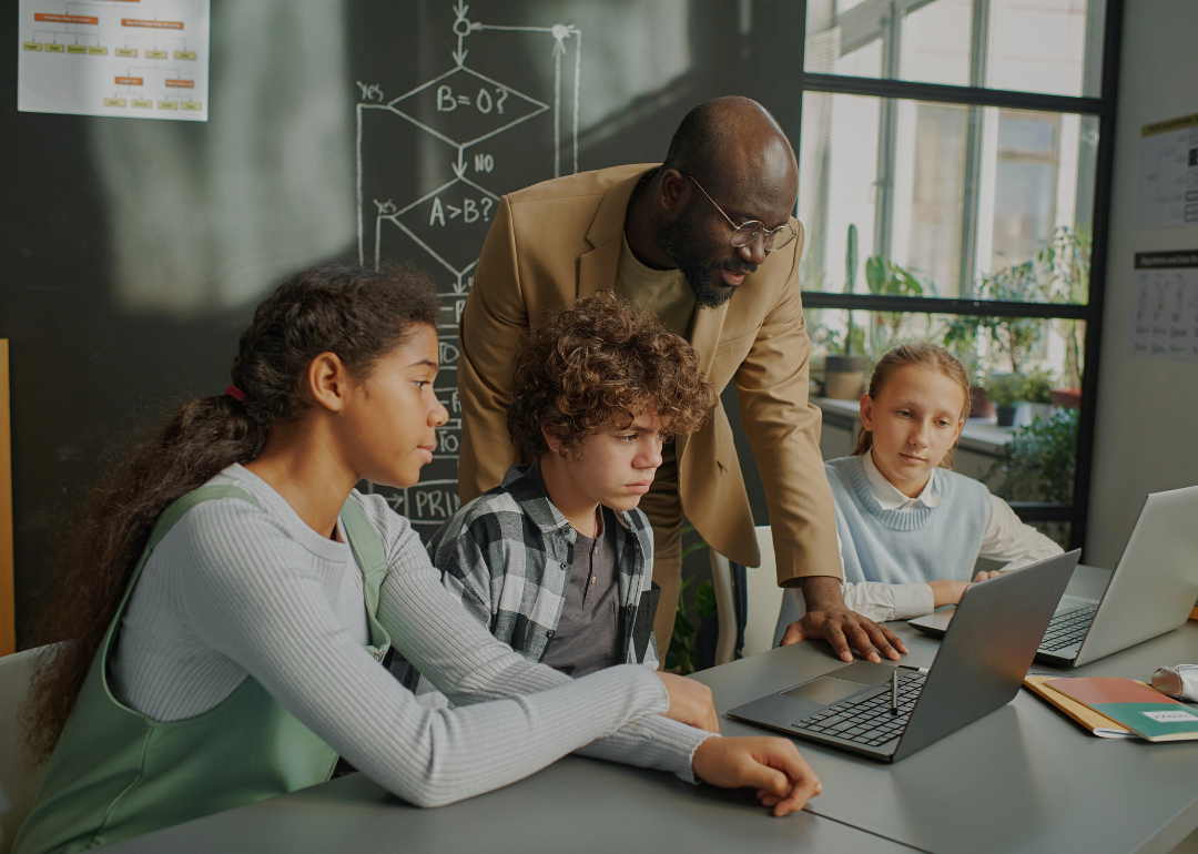 A young Black male teacher looking at the computer screen on the desk, with two girls and a boy focused on the same screen.