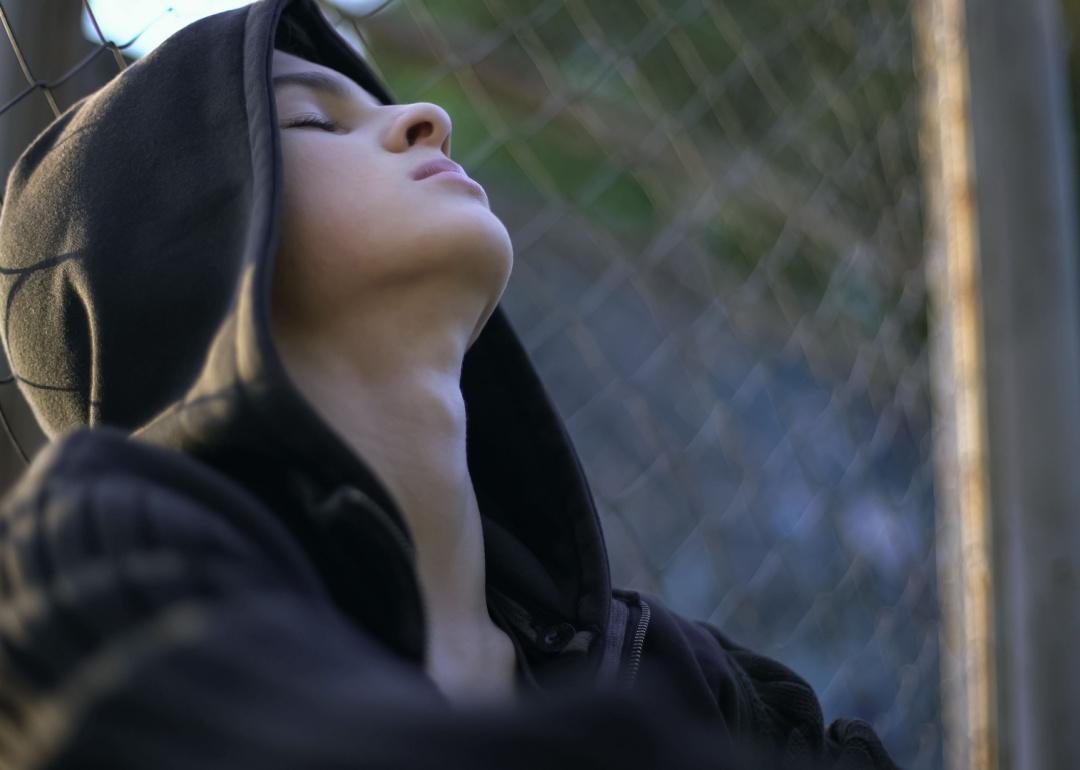Teen wearing a hoodie leaning back on a chain link fence.