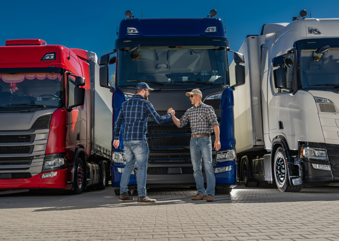 Two men in checked shirts, jeans, and baseball hats shaking hands. There are three truck cabins in the background behind them.