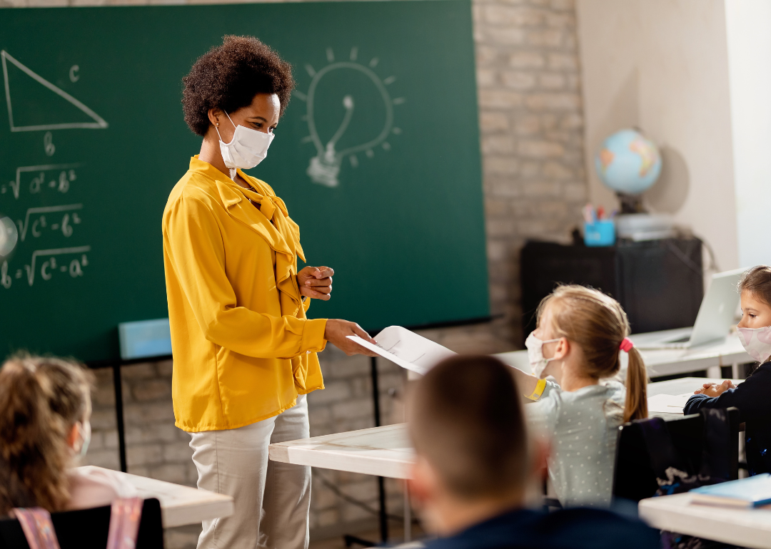 A teacher wearing a mask handing out papers to a class