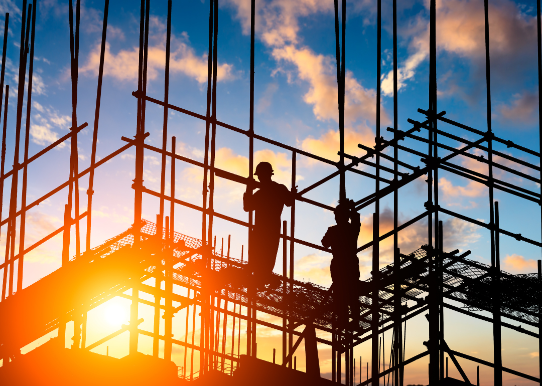 Workers stand on scaffolding in a construction site with an orange sunset in the background.
