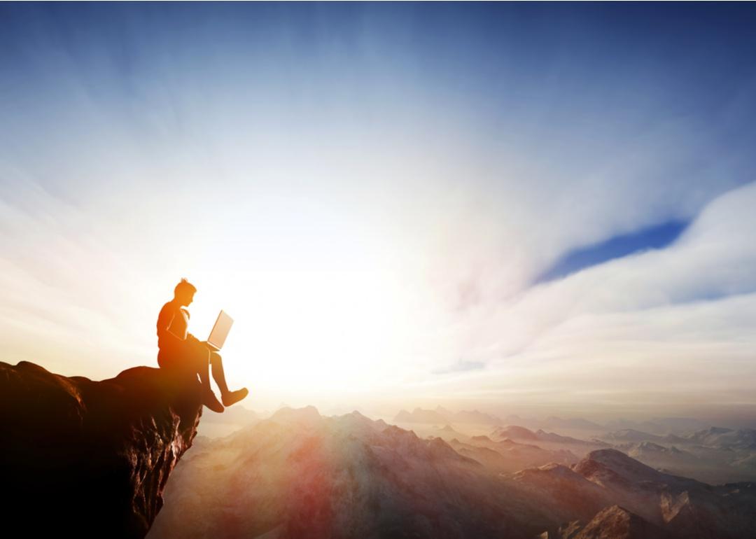 Man sitting on a cliff working on a laptop.