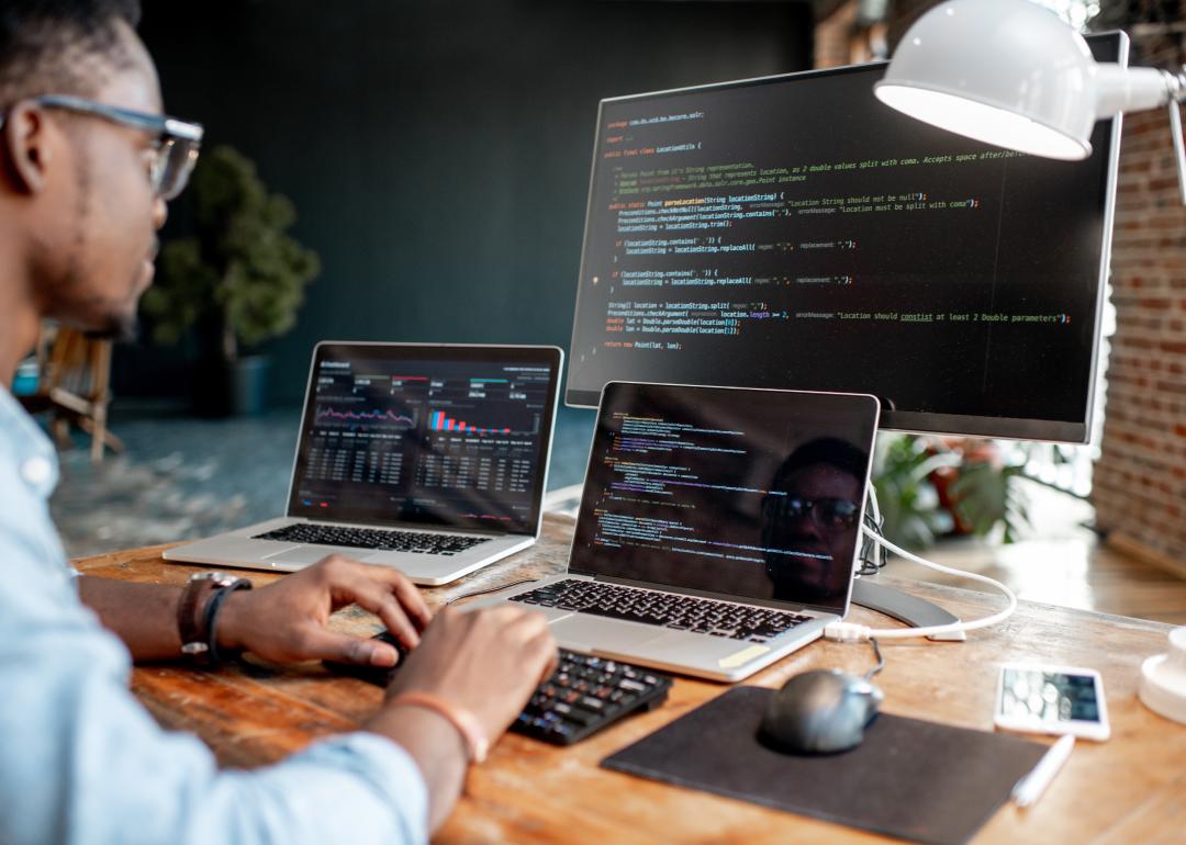 Young male programmer writing code with three monitors on table.